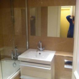 ART.CRAFT. DESIGN.CONSTRUCTIONS - Umywalka z szafką w jasnej łazience wykończonej beżowymi płytkami, z widoczną wanną za szklanym parawanem i odbiciem fotografa w lustrze.