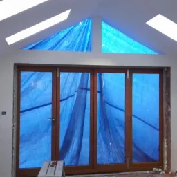 Wnętrze w trakcie prac wykończeniowych: ściany po gładzi, drewniane okno tarasowe zasłonięte niebieską folią ochronną, widoczne ślady po instalacjach elektrycznych i schodki malarskie.