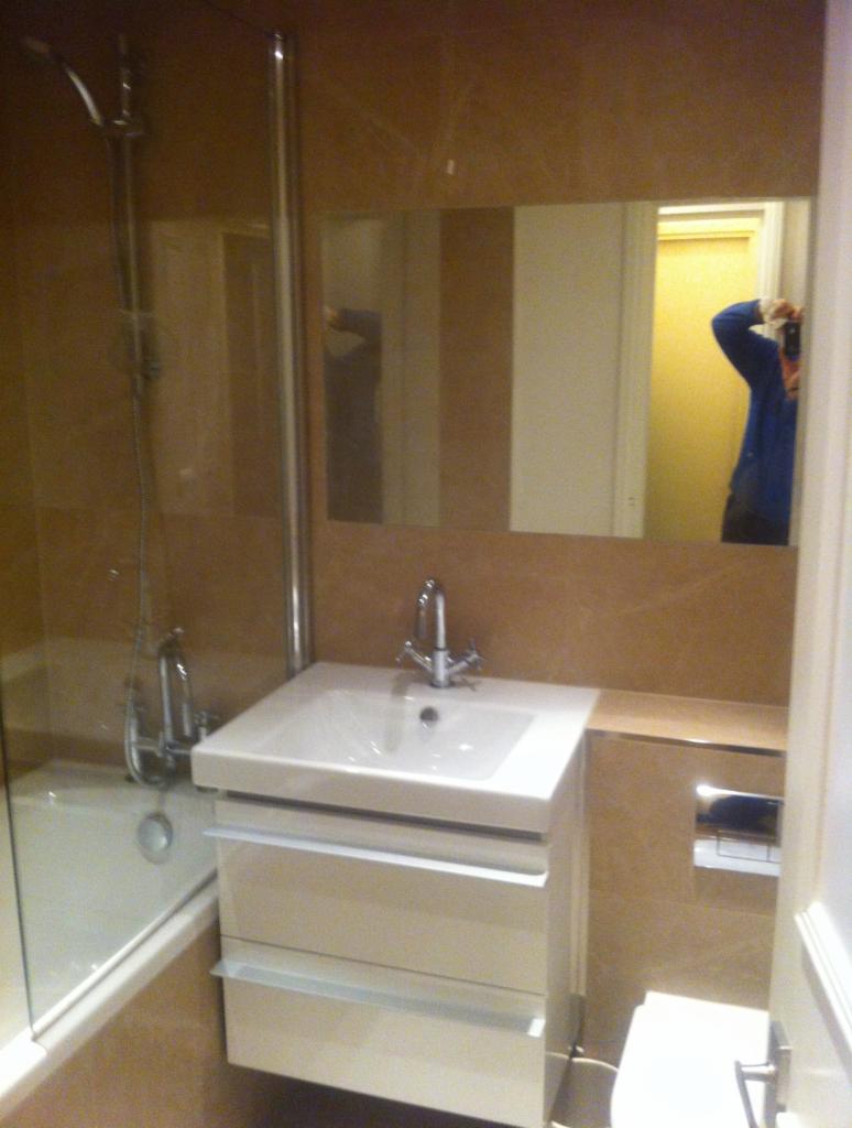 Umywalka z szafką w jasnej łazience wykończonej beżowymi płytkami, z widoczną wanną za szklanym parawanem i odbiciem fotografa w lustrze.