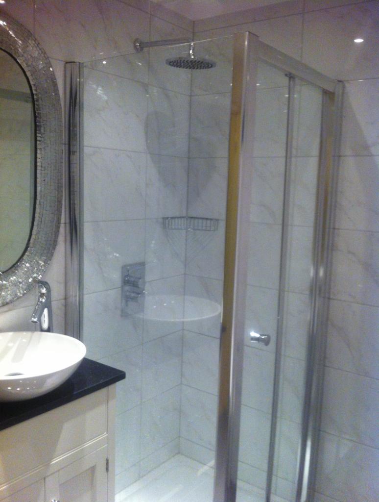 Umywalka z czarnym blatem i owalnym lustrem obok kabiny prysznicowej z deszczownicą, wykończonej białymi płytkami imitującymi marmur.