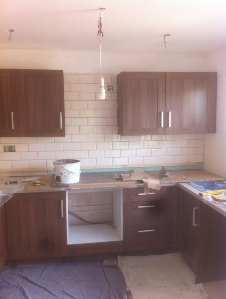 Montaż brązowych szafek kuchennych z białymi uchwytami i białych płytek na ścianie, widoczny biały pojemnik z farbą i narzędzia na blacie, podłoga zabezpieczona folią ochronną, surowa żarówka zwisa...