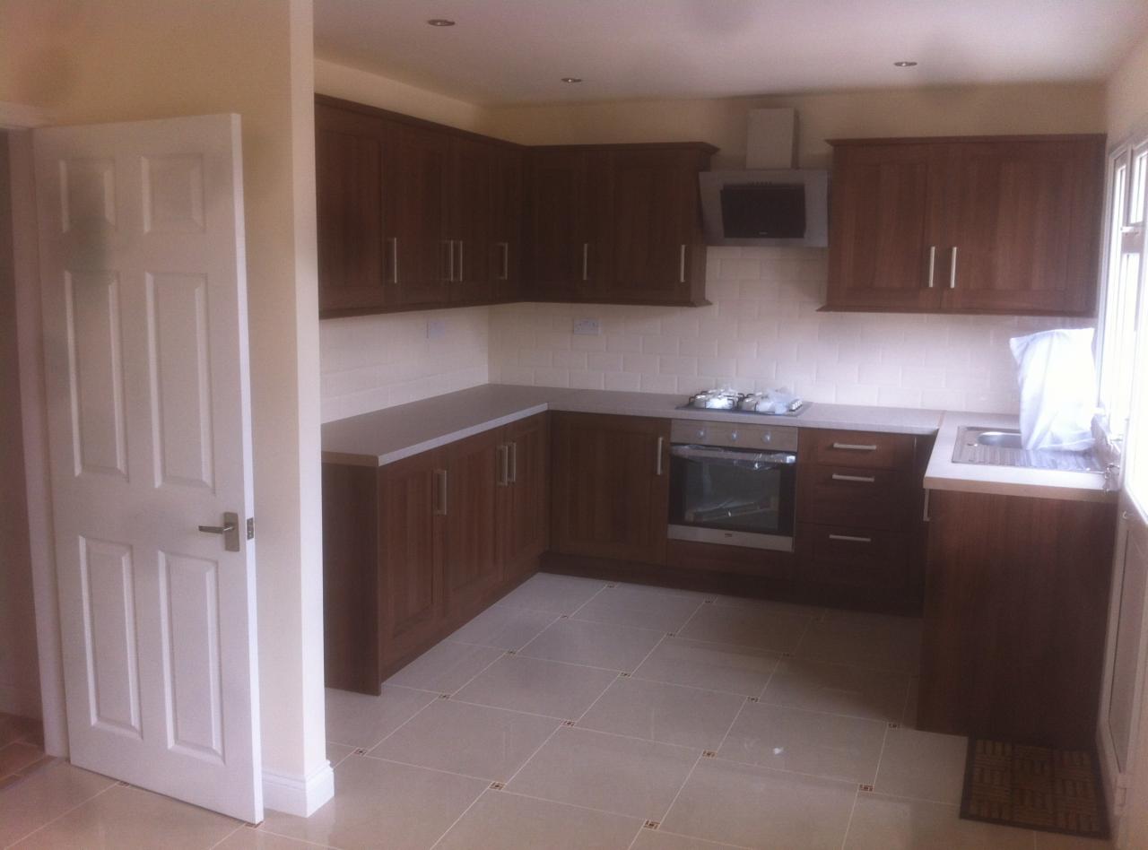 Nowa kuchnia z brązowymi szafkami, jasnymi blatami i beżowymi płytkami podłogowymi, widok z otwartych drzwi.