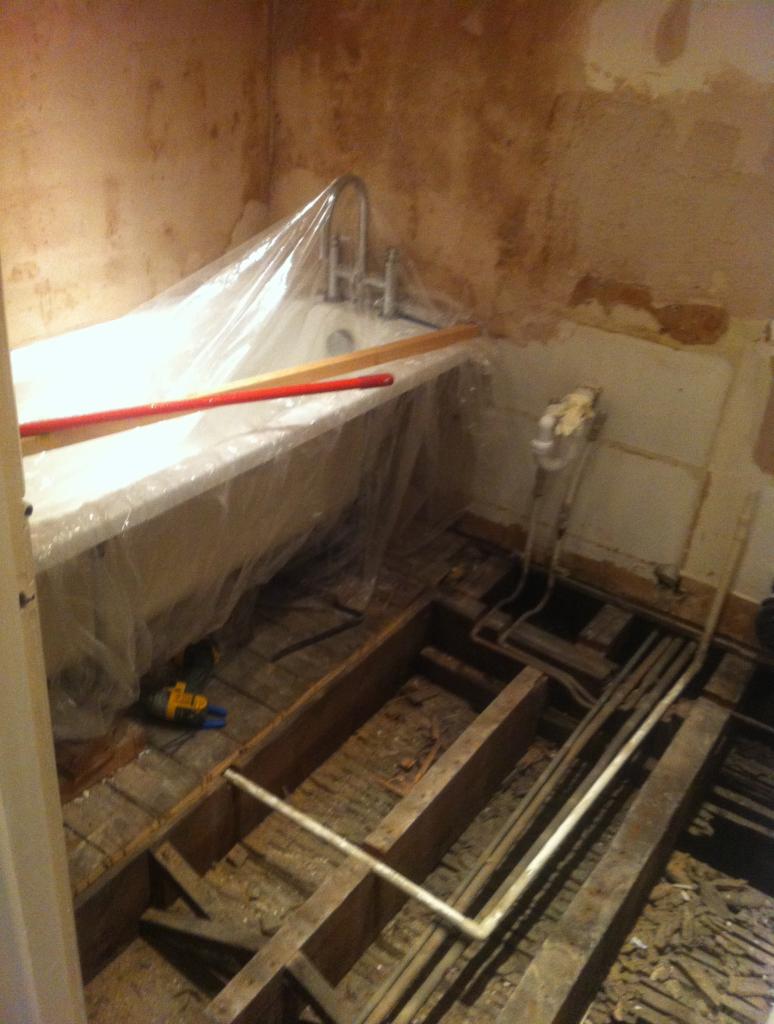 Łazienka w trakcie demontażu podłogi z odkrytymi rurami, wanna zabezpieczona folią, ściany z widocznymi śladami po starych płytkach.