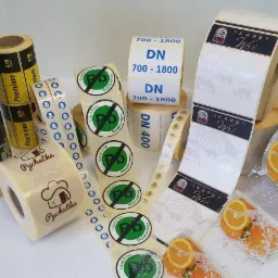 Różnorodne rolki z etykietami i naklejkami: spożywcze, informacyjne (Lead Free), brandingowe (Skarby Wsi, Pychatka), biznesowe (B2B Premium), na białym tle.