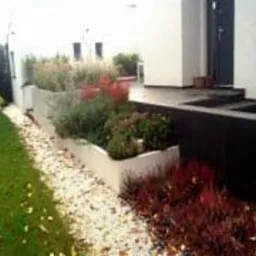 Nowoczesny ogród przydomowy z podwyższonymi rabatami wykończonymi jasnym betonem, wysypany białym grysem ścieżka, zielony trawnik i schody prowadzące do wejścia do domu.