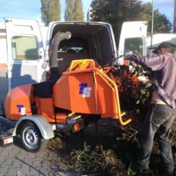 Pracownik w rękawicach wrzuca gałęzie do pomarańczowej rębarki stojącej obok otwartego vana na brukowanym podłożu.