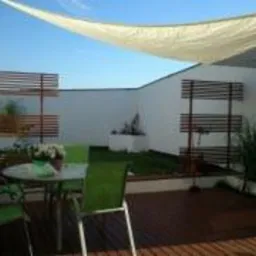 Zewnętrzny taras z drewnianą podłogą, okrągłym stołem i zielonymi krzesłami, zacieniony białym żaglem przeciwsłonecznym. W tle biały mur z elementami dekoracyjnymi z drewna i roślinnością.