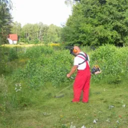 Mężczyzna w czerwonym kombinezonie i ochronnikach słuchu kosi trawę przy użyciu podkaszarki spalinowej na działce z bujną roślinnością i drzewami w tle.
