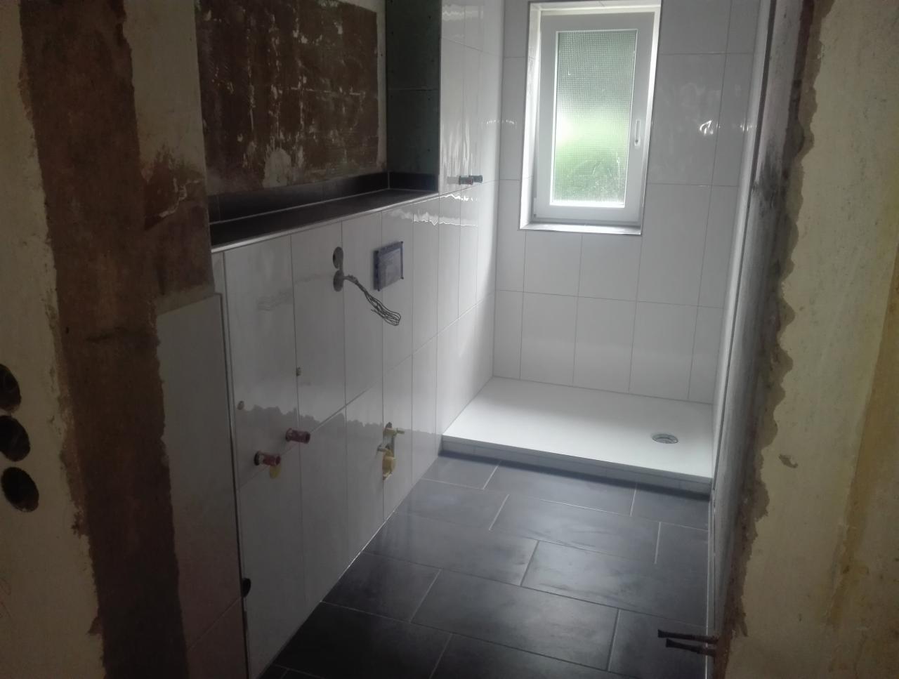 Niedokończona łazienka z widocznymi instalacjami hydraulicznymi i elektrycznymi: białe płytki ścienne, czarna podłoga, niezabudowana ściana z otworami na gniazdka, okno nad brodzikiem.