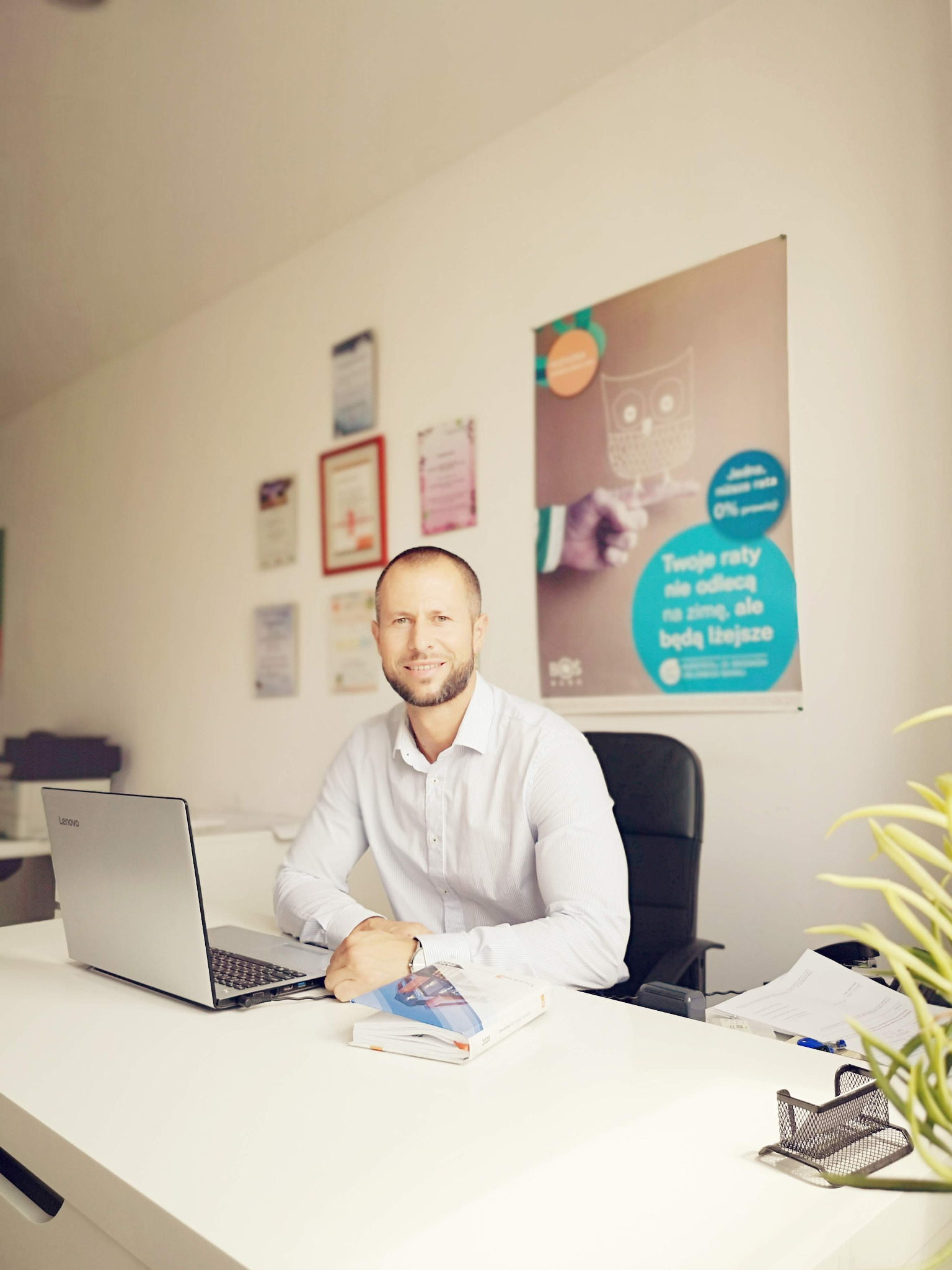 Uśmiechnięty doradca w jasnym biurze siedzi za biurkiem z laptopem Lenovo, na ścianie wisi plakat informujący o ofercie z ratą 0% i rysunkiem sowy, widoczne także certyfikaty i dokumenty.