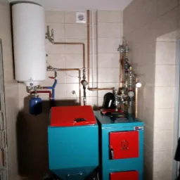 Nowoczesna kotłownia z piecem na paliwo stałe, zasobnikiem ciepłej wody i instalacją z miedzianych rur w pomieszczeniu wyłożonym płytkami ceramicznymi.