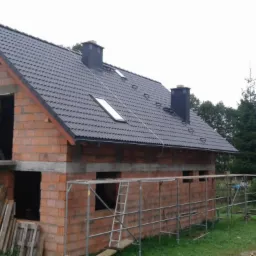 Surowy budynek jednorodzinny w trakcie budowy, z ciemnoszarą blachodachówką, kominami i oknami dachowymi, widoczne rusztowania i materiały budowlane.