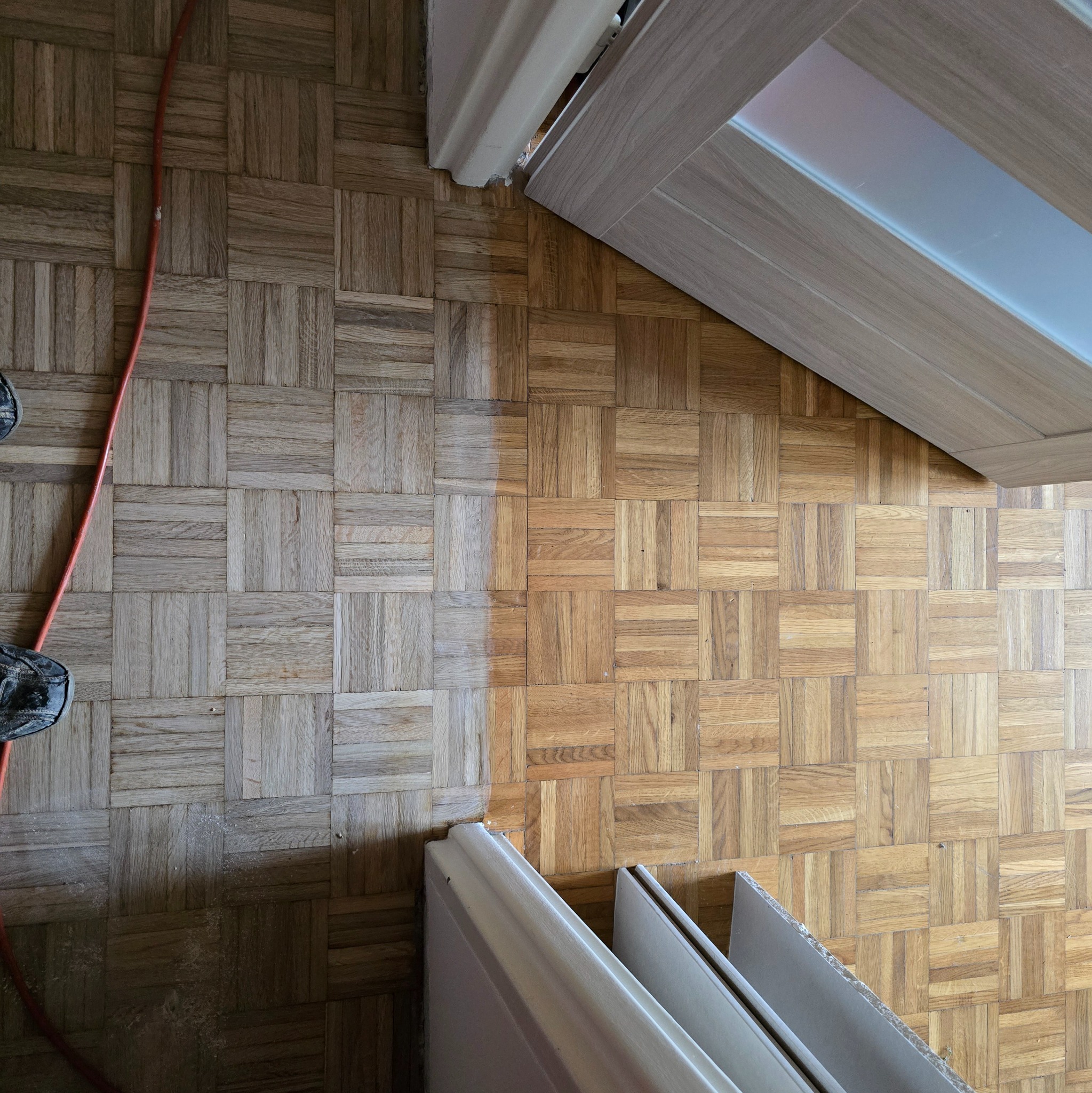 Parkiet w kwadraty, częściowo wycyklinowany, z widocznym kablem i fragmentem buta pracownika, listwy przypodłogowe czekają na montaż.