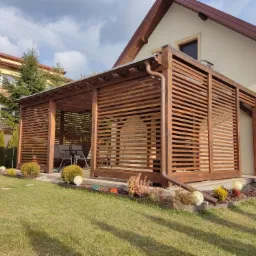 Nowoczesna, drewniana konstrukcja z poziomych listew, przylegająca do jasnego domu z brązowym dachem, otoczona zielonym trawnikiem i ozdobnymi krzewami