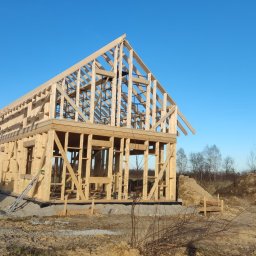 MKWOODBROS Sp. z o.o. - Surowa konstrukcja drewniana domu w trakcie budowy, widoczny szkielet ścian i dachu na tle błękitnego nieba, z przodu widoczny teren budowy i zaparkowany samochód dostawczy.