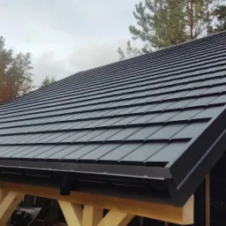 Czarny dach z blachodachówki modułowej na drewnianej konstrukcji altany lub wiaty, widoczne rynny i drzewa w tle.