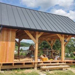 MKWOODBROS Sp. z o.o. - Wola Rafałowska: Budowa drewnianej altany z szarym dachem blaszanym, widoczne drewniane elementy konstrukcyjne, narzędzia i materiały budowlane na placu budowy.