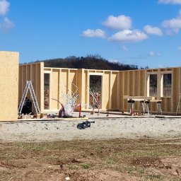 MKWOODBROS Sp. z o.o. - Wznoszenie konstrukcji modułowego domu z drewnianych paneli na fundamencie, widoczny pracownik w otworze okiennym, narzędzia i agregat prądotwórczy w tle.