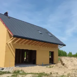 Wznoszony dom z drewnianą konstrukcją, pokryty blachą na rąbek stojący z oknami połaciowymi, pracownik na dachu, widoczna warstwa izolacji pod blachą, na pierwszym planie hałda piasku