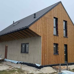 MKWOODBROS Sp. z o.o. - Surowy stan domu z drewnianą elewacją i czarnym dachem blaszanym, widoczne rusztowanie i materiały budowlane wokół, okna z czarnymi ramami.