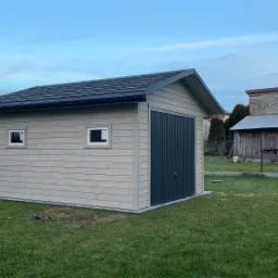 Nowoczesny, drewniany garaż z dwoma małymi oknami i ciemnymi drzwiami segmentowymi, na tle zielonego trawnika i pochmurnego nieba.