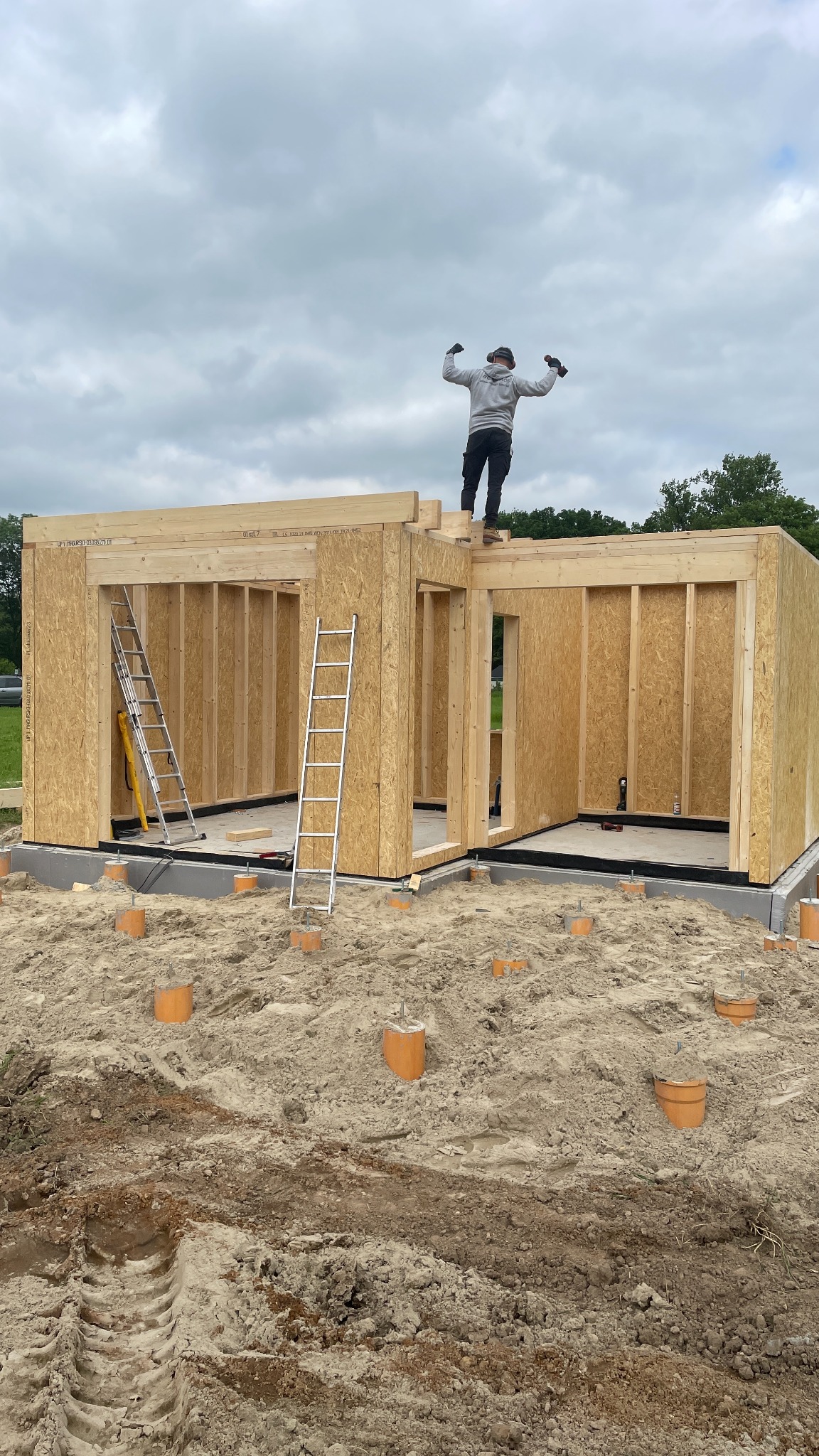 Wznoszenie konstrukcji domu z drewnianych paneli OSB na fundamencie, pracownik na dachu z podniesionymi rękami, widoczne dwie drabiny i pomarańczowe rury wentylacyjne w gruncie.