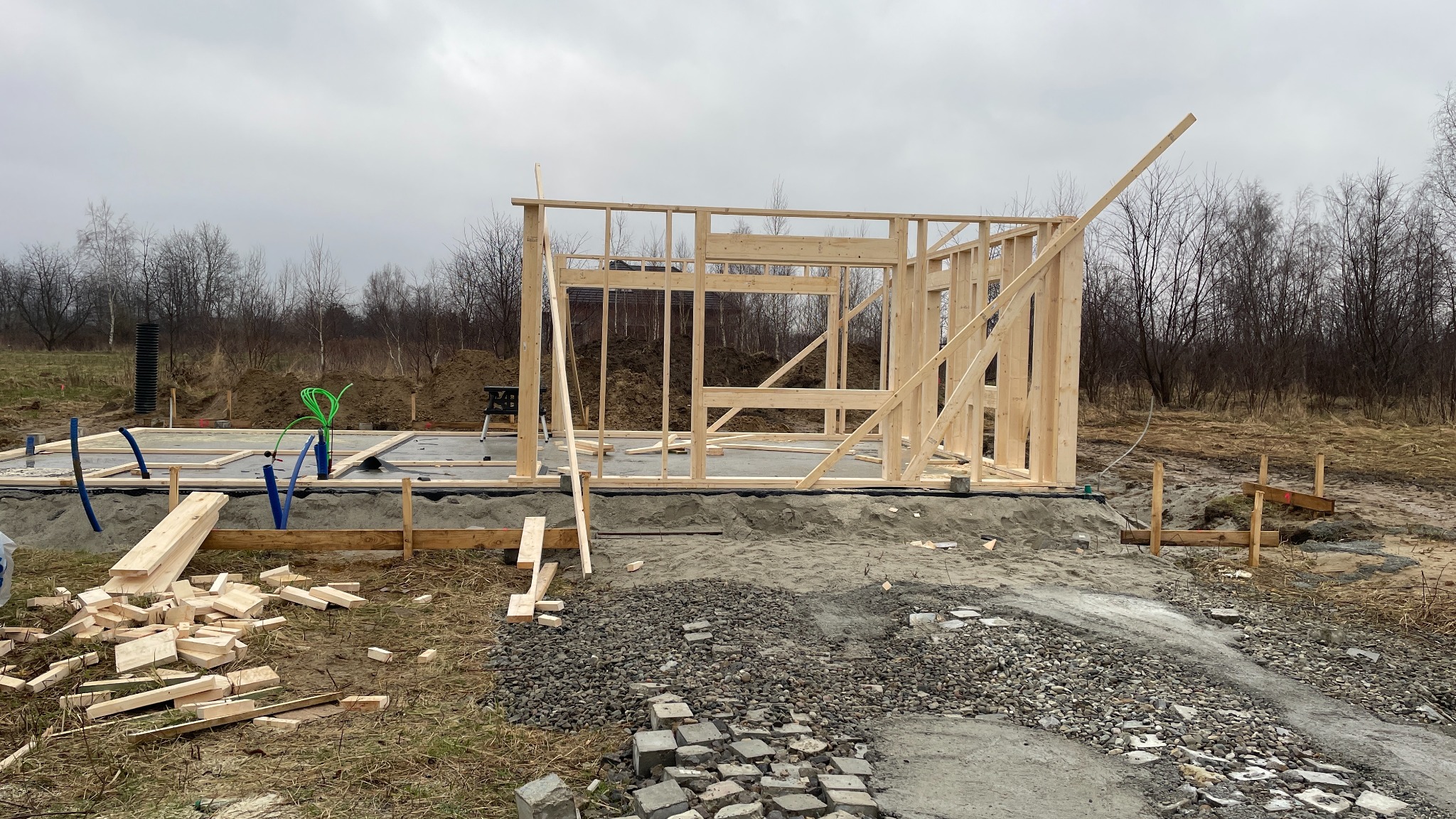 Wznoszenie drewnianej konstrukcji szkieletowej na betonowej płycie fundamentowej, widoczne niebieskie rury instalacyjne i porozrzucane elementy drewniane.