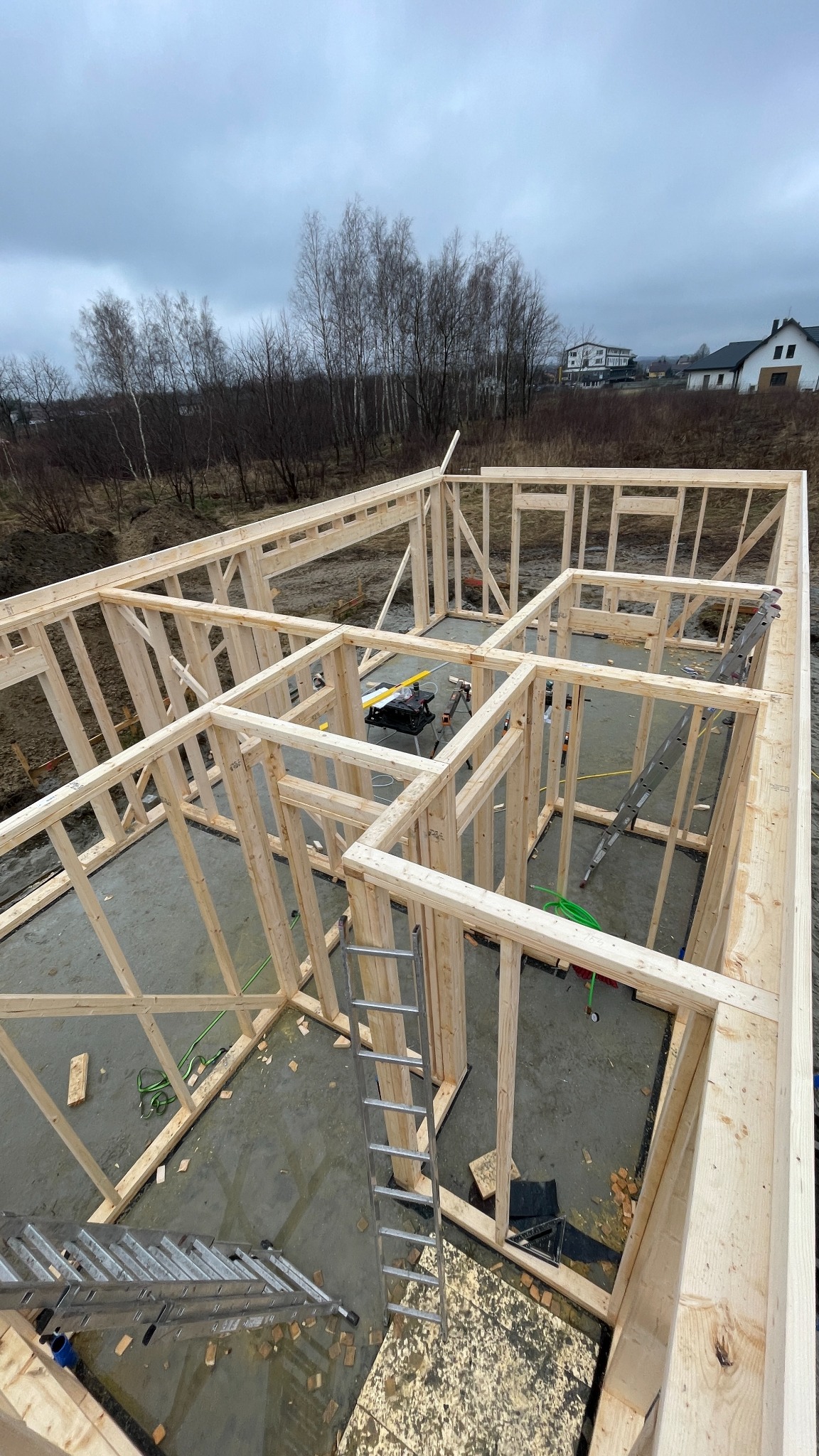 Wznoszenie szkieletu drewnianego domu na betonowej płycie fundamentowej, widoczne drabiny, narzędzia i elementy konstrukcyjne, w tle drzewa i zabudowania.