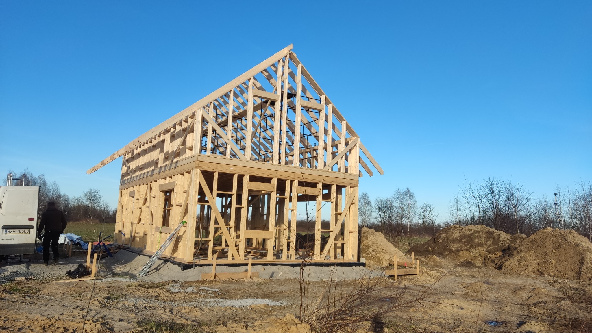 Surowa konstrukcja drewniana domu w trakcie budowy, widoczny szkielet ścian i dachu na tle błękitnego nieba, z przodu widoczny teren budowy i zaparkowany samochód dostawczy.