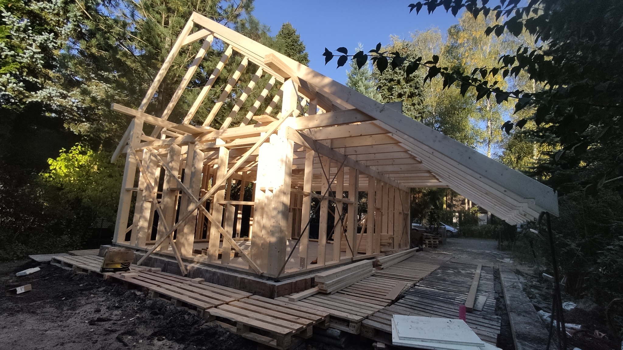 Konstrukcja szkieletowa domu w trakcie budowy, widoczne drewniane elementy nośne i zadaszenie, otoczenie leśne.