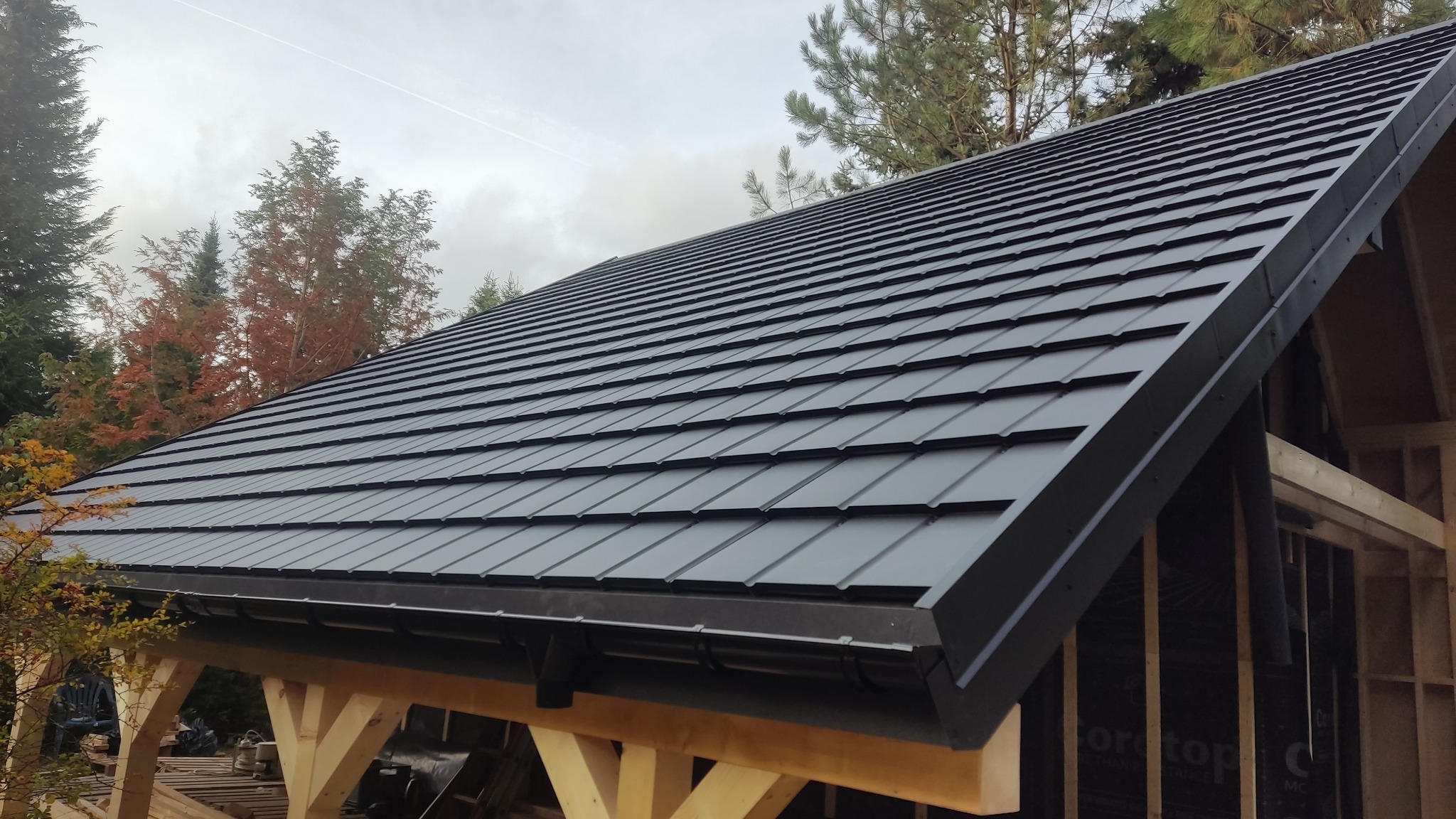 Czarny dach z blachodachówki modułowej na drewnianej konstrukcji altany lub wiaty, widoczne rynny i drzewa w tle.