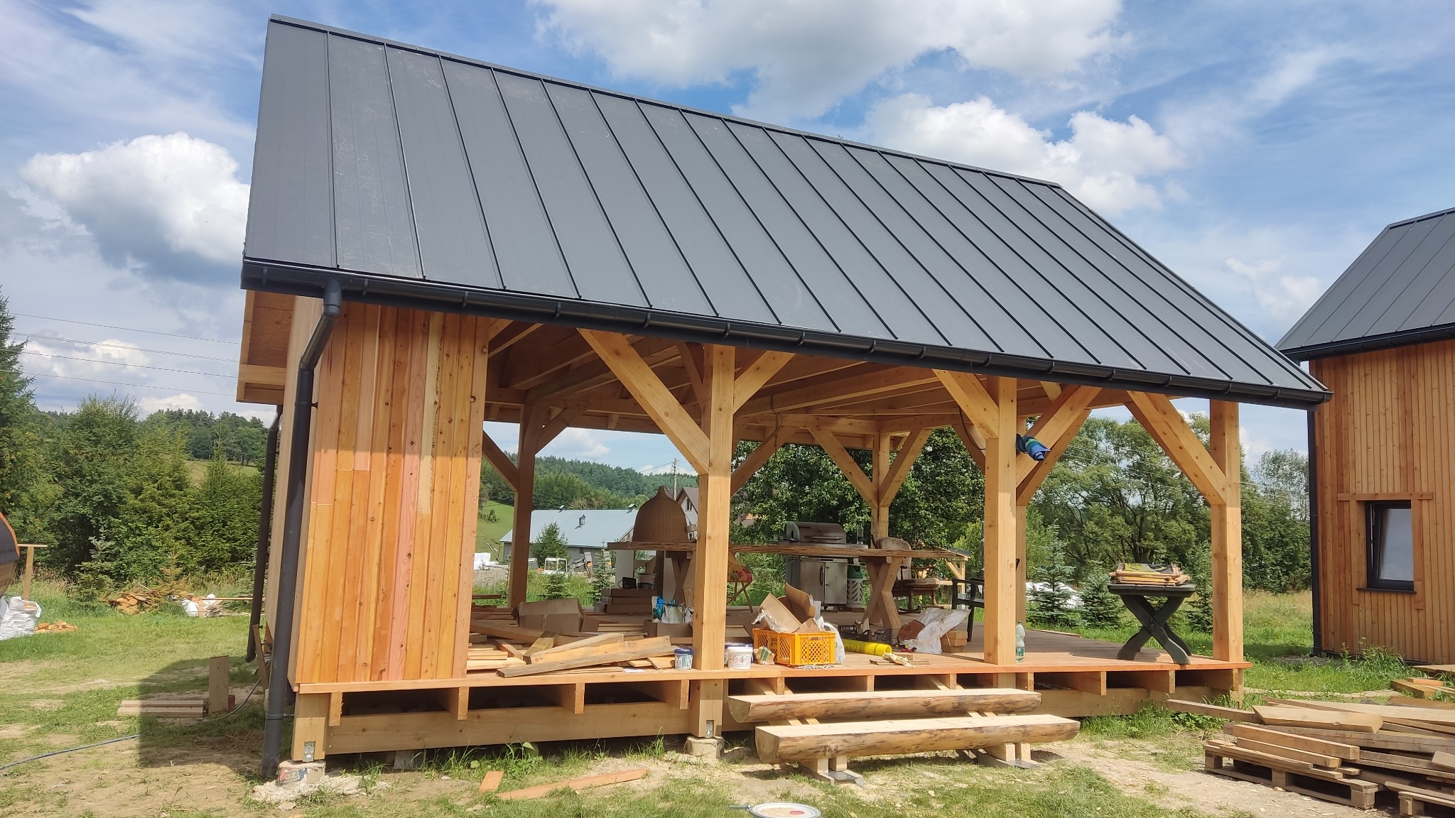 Wola Rafałowska: Budowa drewnianej altany z szarym dachem blaszanym, widoczne drewniane elementy konstrukcyjne, narzędzia i materiały budowlane na placu budowy.