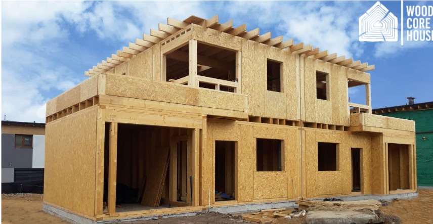 Surowa konstrukcja dwukondygnacyjnego budynku z drewnianych paneli OSB, widoczne otwory okienne i zarys dachu, na górze logo firmy Wood Core House.