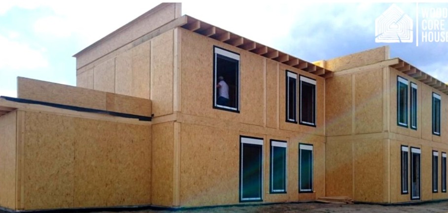 Wznoszony dwukondygnacyjny budynek z płyt drewnopochodnych z zamontowanymi oknami, widoczna osoba w oknie na piętrze, logo producenta w prawym górnym rogu.