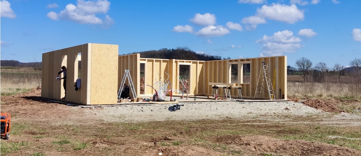 Wznoszenie konstrukcji modułowego domu z drewnianych paneli na fundamencie, widoczny pracownik w otworze okiennym, narzędzia i agregat prądotwórczy w tle.