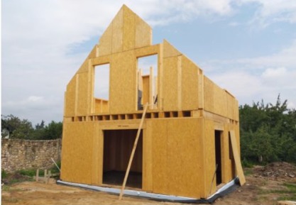 Wznoszenie dwukondygnacyjnej konstrukcji budynku z drewnianych paneli OSB, widoczne otwory okienne i drzwiowe, na zewnątrz kamienny murek i zieleń