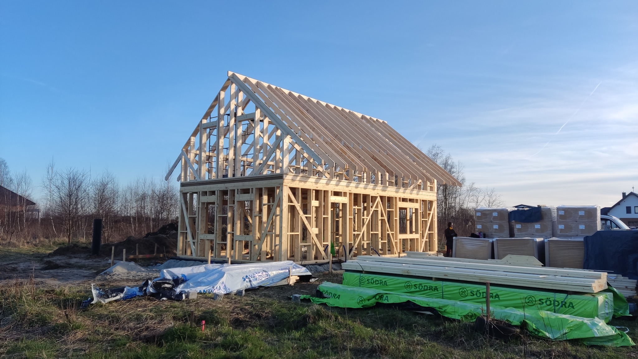 Wznoszenie drewnianej konstrukcji domu w technologii szkieletowej, widoczne elementy ścian i dachu, na pierwszym planie materiały budowlane zabezpieczone folią