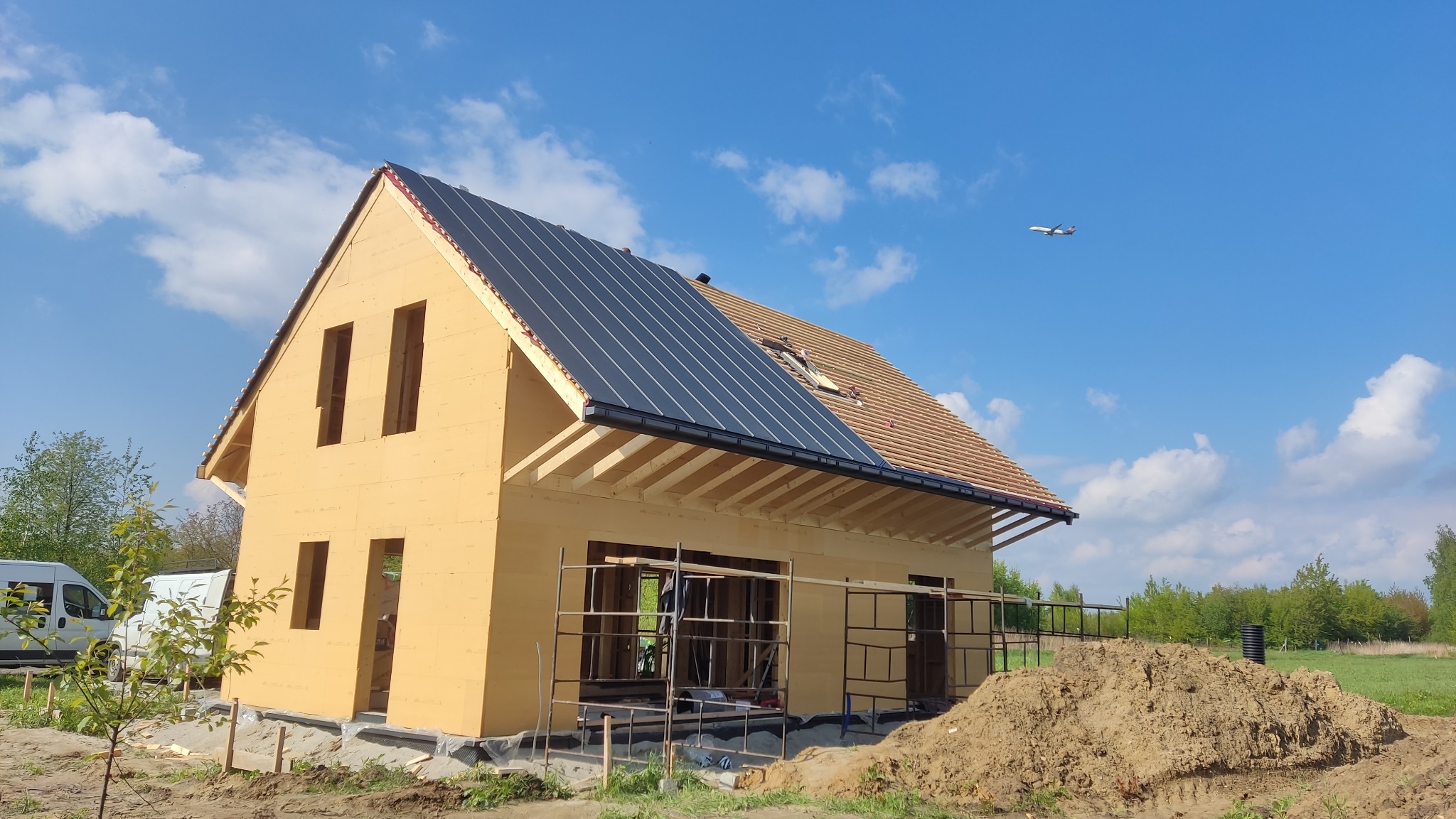 Surowy stan domu w konstrukcji szkieletowej z częściowo ukończonym dachem, na którym widać dwa rodzaje pokrycia oraz przelatujący samolot na tle błękitnego nieba.