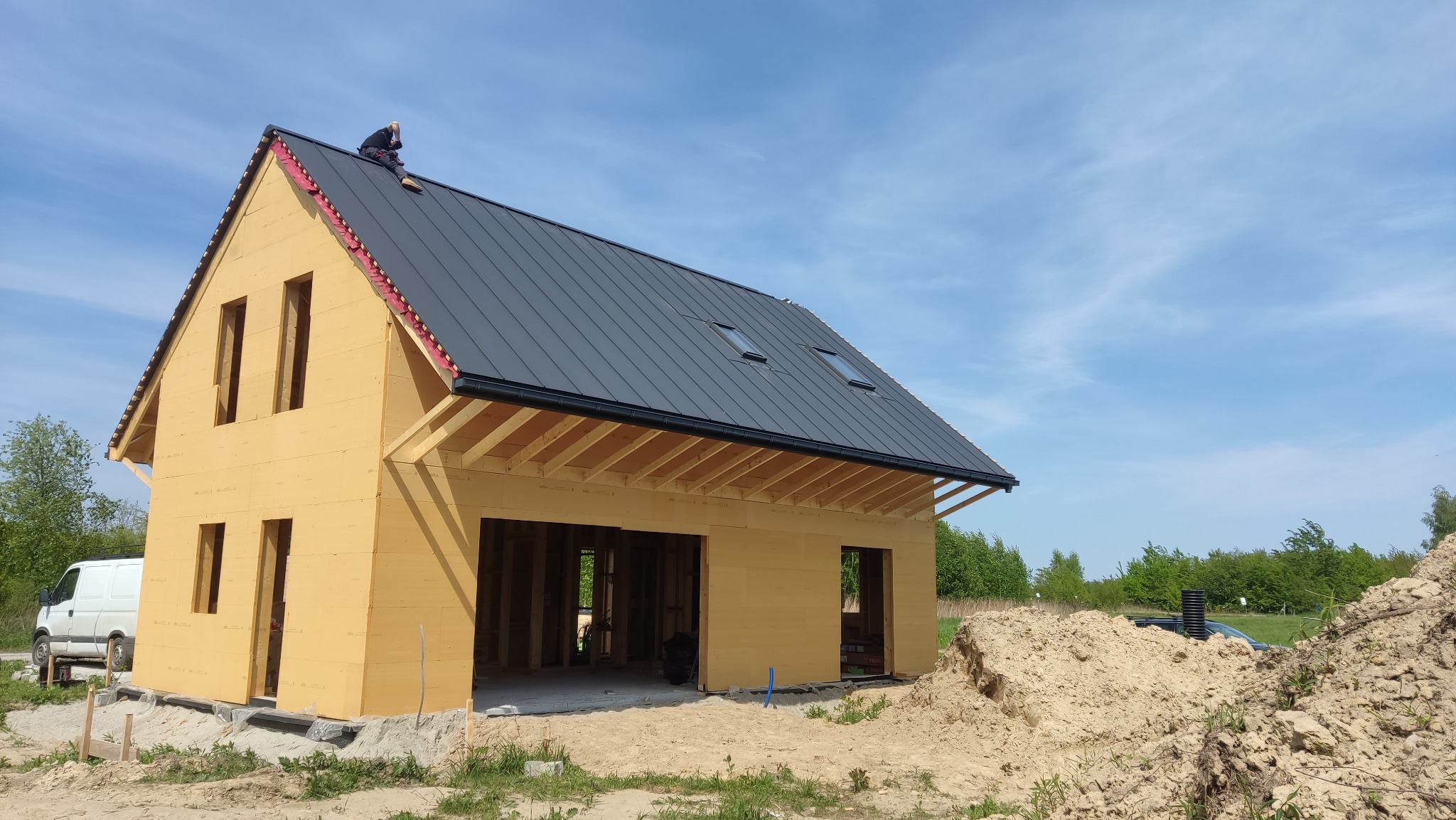 Wznoszony dom z drewnianą konstrukcją, pokryty blachą na rąbek stojący z oknami połaciowymi, pracownik na dachu, widoczna warstwa izolacji pod blachą, na pierwszym planie hałda piasku
