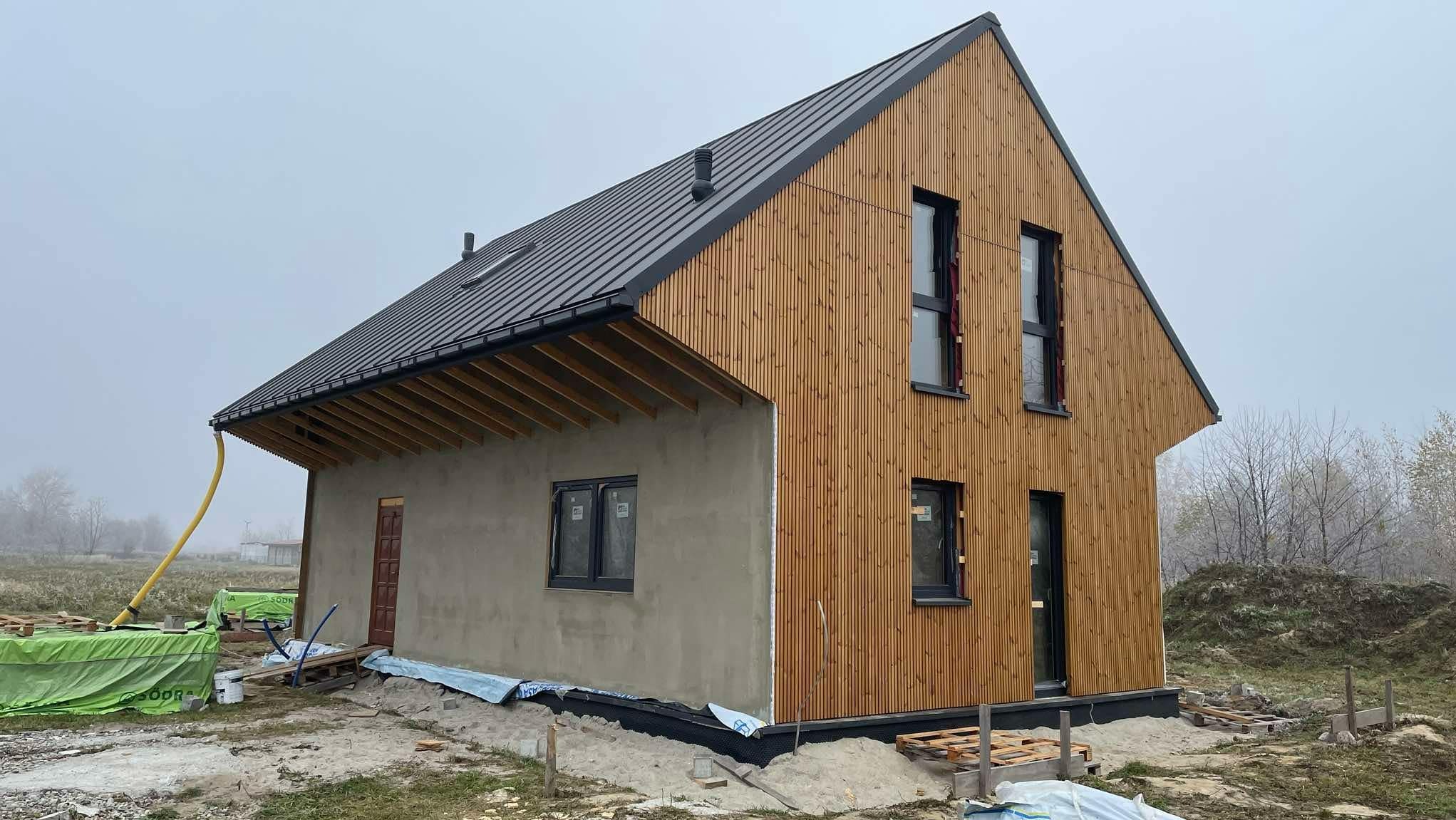 Surowy stan domu z drewnianą elewacją i czarnym dachem blaszanym, widoczne rusztowanie i materiały budowlane wokół, okna z czarnymi ramami.