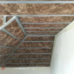 Skośny sufit poddasza w trakcie izolacji wełną mineralną, widoczne metalowe profile konstrukcyjne i fragment ściany z taśmą uszczelniającą.