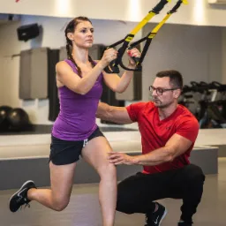 Kobieta ćwiczy z użyciem taśm TRX, wspierana przez mężczyznę w okularach, w tle lustro i sprzęt do ćwiczeń.