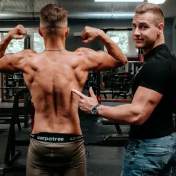 Mężczyzna prezentuje umięśnione plecy w siłowni, trener wskazuje palcem na mięśnie, w tle sprzęt do ćwiczeń.