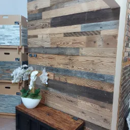 Ściana z desek o zr&oacute;żnicowanej kolorystyce i fakturze, po lewej stronie ekspozycja płytek imitujących kamień, na dole drewniana szafka z metalowymi drzwiczkami, a na niej biały wazon z orchideami.