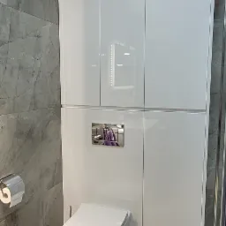 Nowoczesna łazienka z wiszącą toaletą, zabudową szafkową w białym połysku i szarą ścianą imitującą marmur.