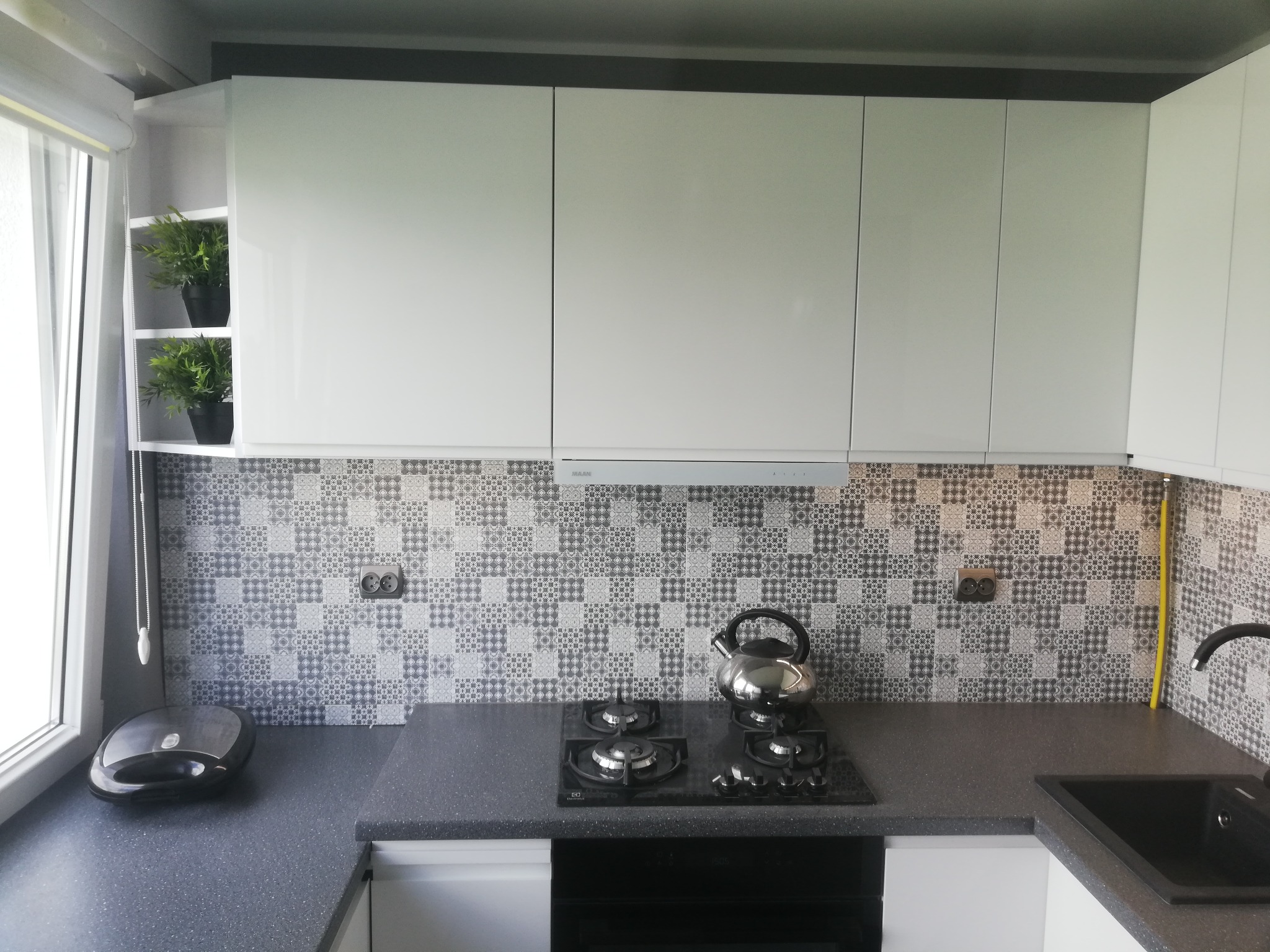 Nowoczesna kuchnia w Grudziądzu z białymi szafkami, grafitowym blatem i mozaikową ścianą nad płytą gazową; na blacie czajnik ze stali nierdzewnej i opiekacz.