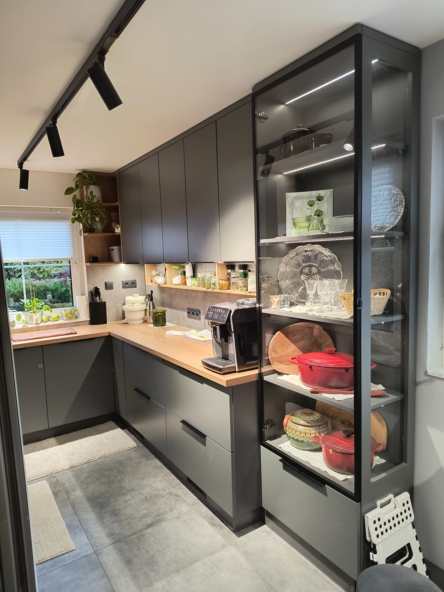 Nowoczesna kuchnia w odcieniach szarości z drewnianym blatem, oświetleniem punktowym i przeszkloną witryną z dekoracyjną zastawą.