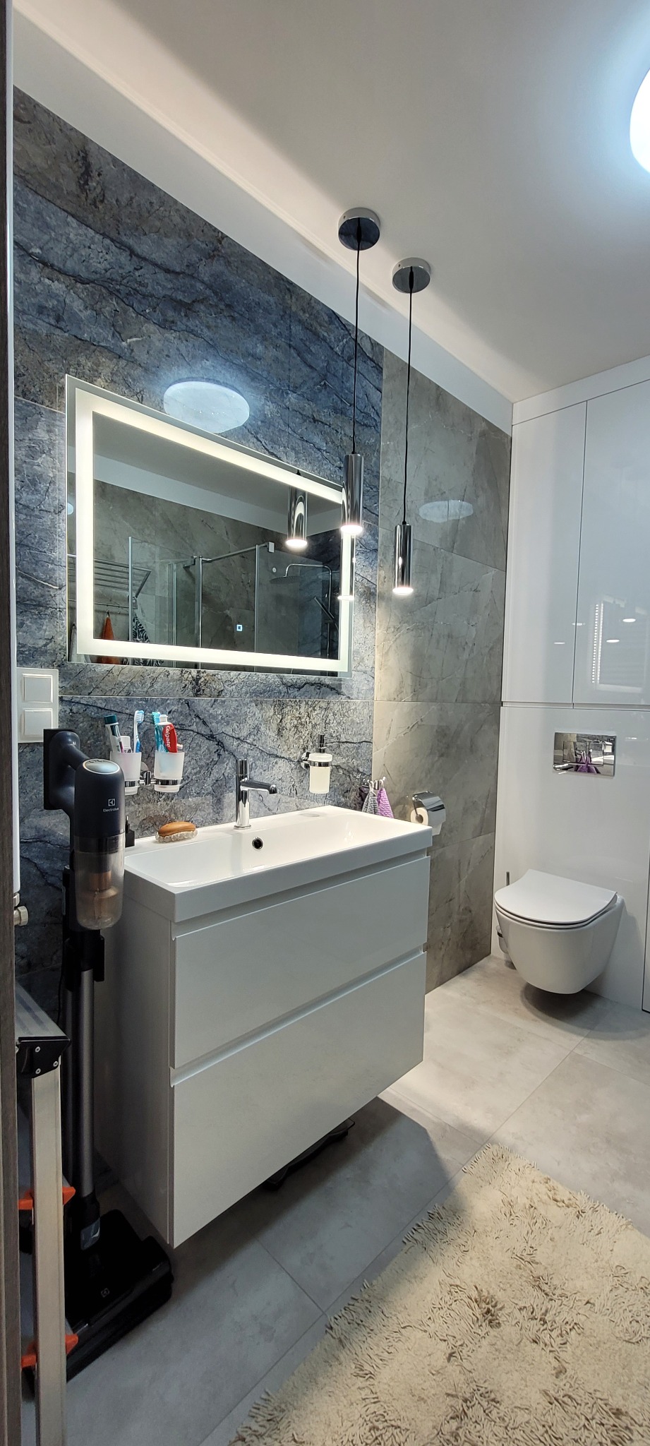 Nowoczesna łazienka z szarą marmurową ścianą, podświetlanym lustrem, białą szafką pod umywalką i wiszącą toaletą, oświetlona wiszącymi lampami.