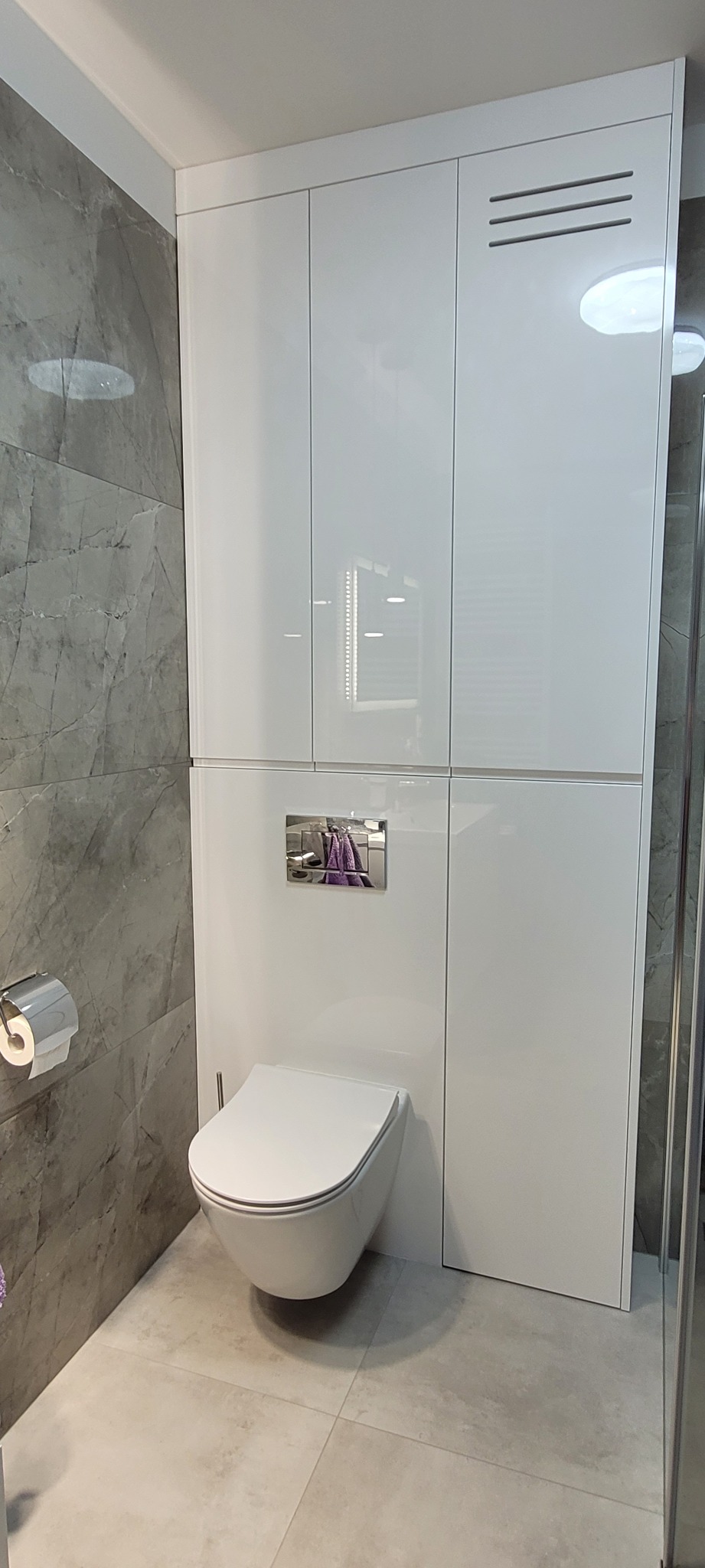 Nowoczesna łazienka z wiszącą toaletą, zabudową szafkową w białym połysku i szarą ścianą imitującą marmur.