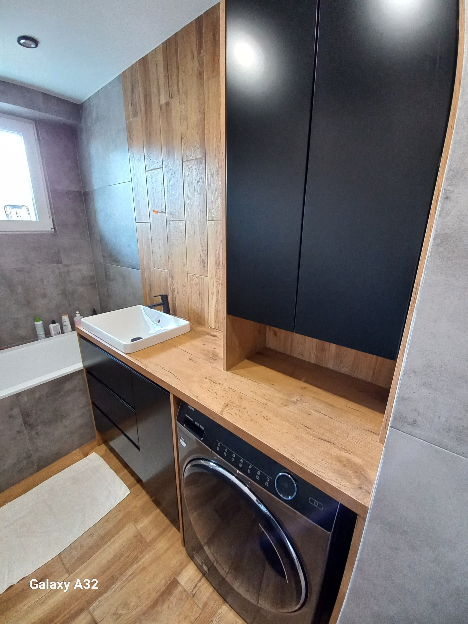Nowoczesna łazienka z umywalką nablatową, czarnymi szafkami pod umywalką i pralką zabudowaną pod blatem z drewnopodobnym wykończeniem, oraz płytkami imitującymi drewno na ścianie.
